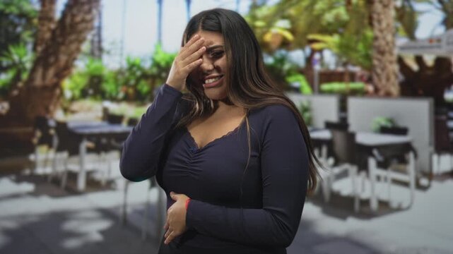 Woman covering face with hand, visible cleavage and hand on stomach, standing by outdoor cafe tables and palm trees on street; embarrassment.