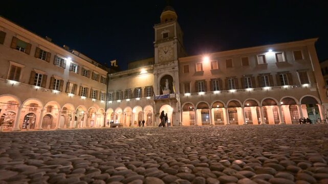 Modena, italy, april 11, 2026. Low angle view of the cobblestone pavement in piazza grande, modena. Historical buildings and clock tower illuminated at night