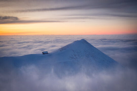 Lone cabin on snowy peak above sea of clouds at sunset
