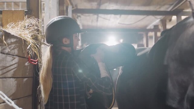 Helmeted rider adjusting saddle and tack. Tandem education moment with assistant in busy barn aisle, checking girth and bridle, focused teamwork, warm backlight and concentrated effort.