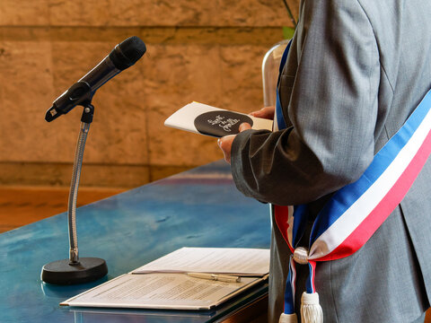 Officiant de mariage civil avec &eacute;charpe tricolore lisant un document au micro en mairie
