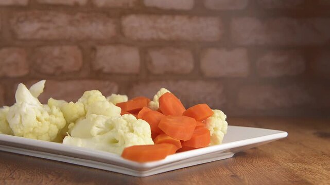 Steamed vegetables, carrots and cauliflower on a rectangular white plate to accompany meat, chicken or fish, on a rustic wooden table, with a brick wall in the background, ready to be seasoned
