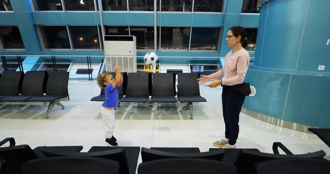 Toddler and mom play with soft ball in empty airport terminal during flight delay. Young child throws ball, and mother passes it back. Son joyfully hits it with both hands, like in volleyball