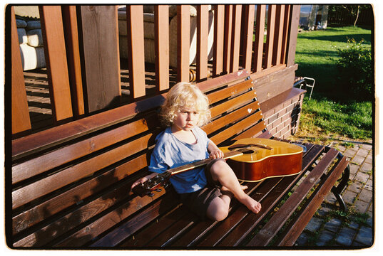 A blond boy sits on a bench and looks at a guitar