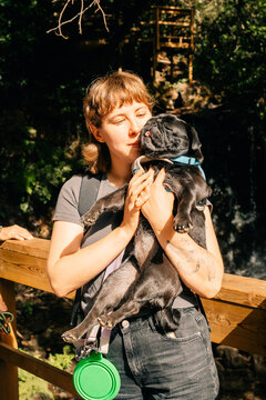 Woman Affectionately Holds Pug Near Waterfall in Sunny Park