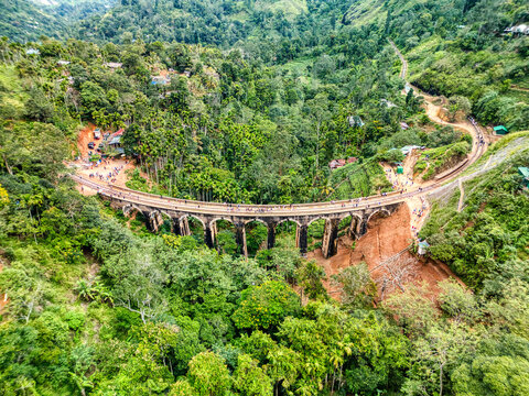 Aerial view of Nine Arch Bridge with people walking along the stone viaduct surrounded by dense tropical jungle and lush greenery in Demodara, Uva Province, Sri Lanka.