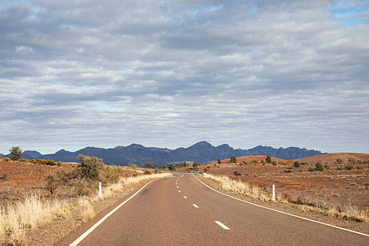 Scenic outback road leading towards rugged mountains under cloudy sky