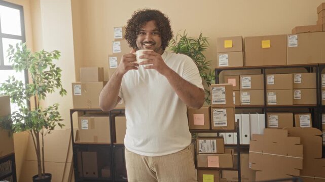 Young man holding a coffee cup and smiling among stacked cardboard boxes and shipping labels in building; contentment.