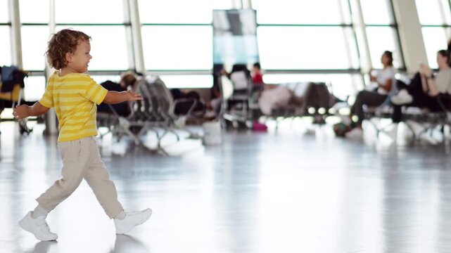 Joyful toddler kicks soft ball across airport departure lounge, spinning around in excitement to celebrate his success. Slow-motion, full-length side view, with blurred passenger lounge on background