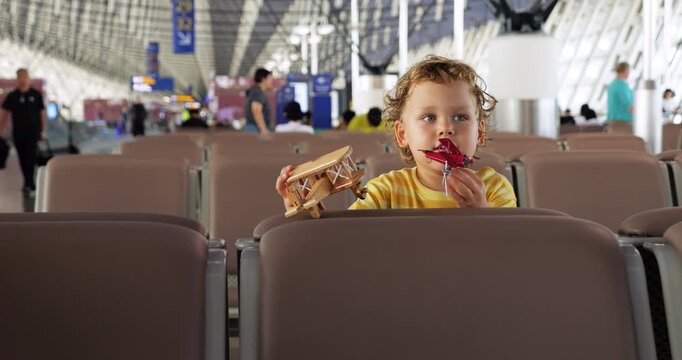 Toddler boy sits calmly in public lounge of large airport, holding two toy airplanes and thoughtfully showing them. Spacious empty boarding gate area of international terminal blurred in background
