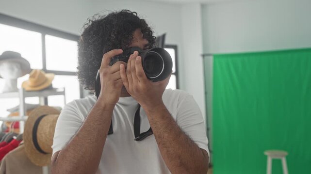 Young indian man photographer holds camera to eye, hands framing lens near green screen and hat rack in studio; focused creativity.