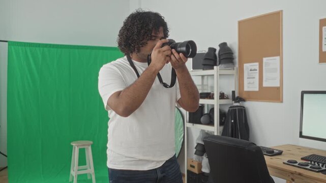 Man holding camera with hands, raises camera to eye surrounded by green screen and studio gear, smiling and adjusting lens; confidence.