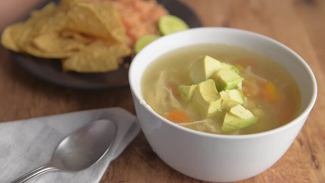 Mexican food (TLALPE&Ntilde;O broth) in a white bowl with vegetables, chicken, avocado and a chipotle chili on top, accompanied by a black plate of totopos with Mexican rice and some pieces of lemon.