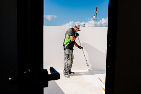 Elderly painter applying white paint to a terrace viewed from behind