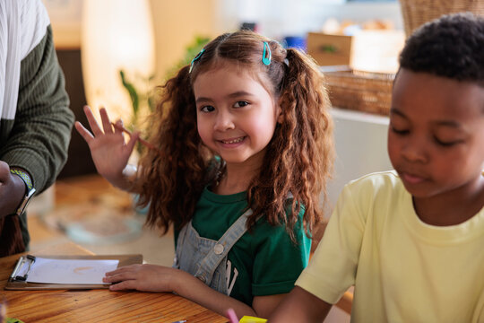 Children drawing at a table, one waving hello.