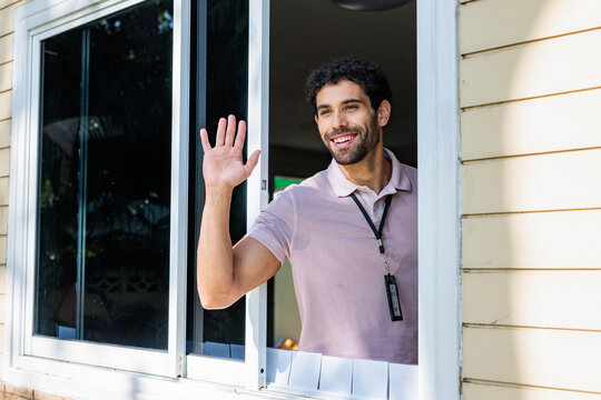 Man waves from window, wearing lanyard and smiling.
