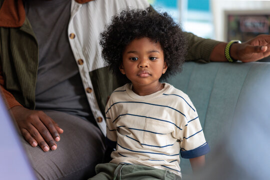 Child with curly hair sits next to adult on couch.