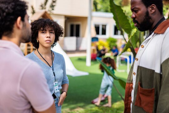 Three adults converse outdoors, children play in the background.