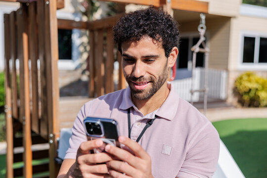 Man smiles while using phone outdoors near playground.