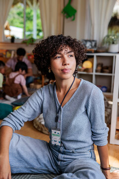 Thoughtful teacher sits with children in background at classroom.