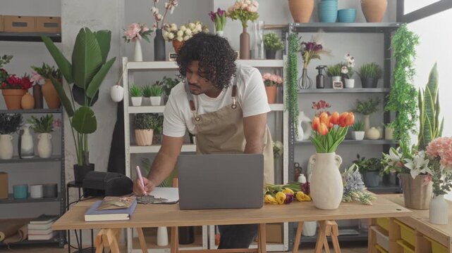 Man florist in apron leans on table with laptop and tulips, hands writing on clipboard in studio; warmth and craft.