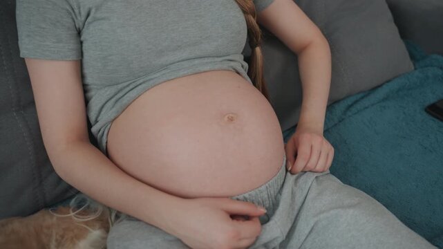 Pregnant white woman examining belly closely, fingers near navel counting kicks, focused expression, braided hair, loungewear, closeup living room shot, natural light, mindful prenatal check.
