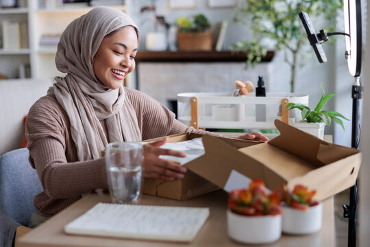Smiling woman in hijab opens box for online video.