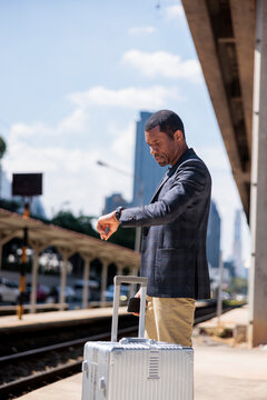 Man with suitcase checks watch at train station.