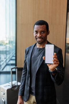 Smiling man shows blank smartphone screen at airport.
