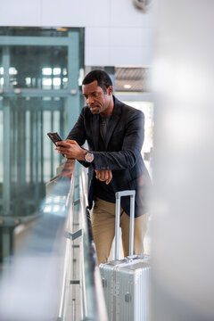 Man with suitcase checks phone at airport railing.