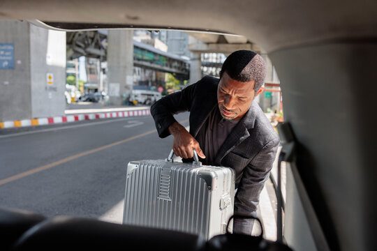 Man loads silver suitcase into car trunk in city.