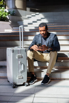 Traveler checks phone while waiting on steps with luggage.