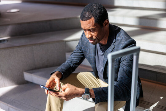 Man on stairs checks phone while waiting with luggage.