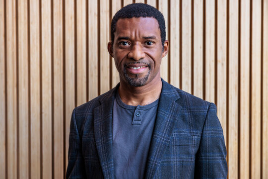 Smiling man in blazer against a wooden wall.