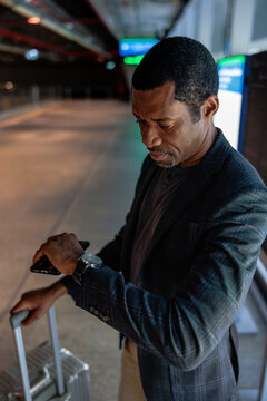 Man checks watch while waiting with luggage at station.