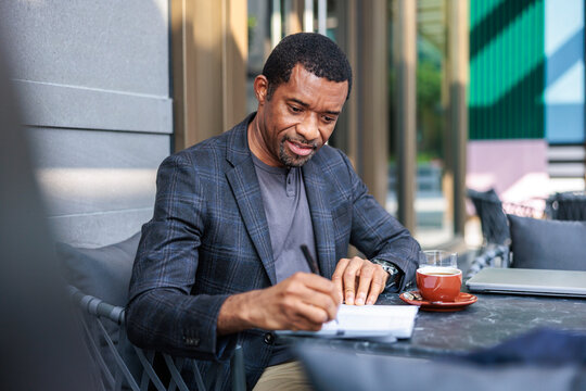 Man writing in notebook with coffee and laptop nearby.