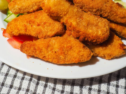 Golden, crispy breaded fried chicken pieces glow under warm light. They rest on a plain white plate and looking freshly cooked. Each piece has a textured, crunchy exterior. Fine goujons ready to eat