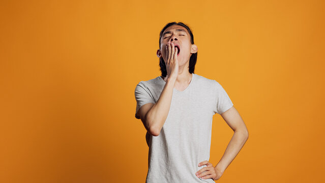 Drained person yawning and being tired in studio, falling asleep and feeling worn out. Exhausted young man being sleepy and carefree over orange background, having confidence on camera.