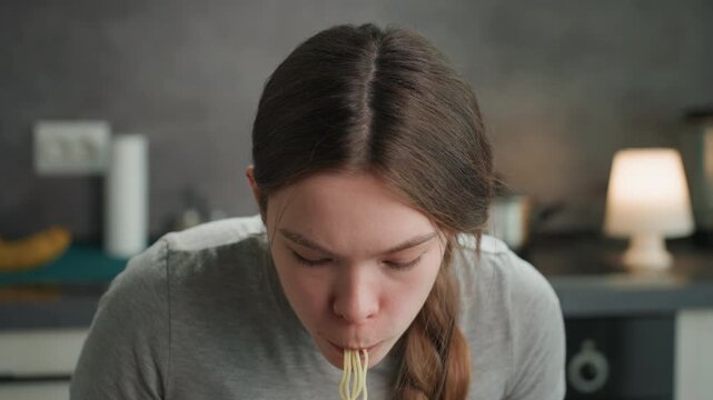 Pregnant woman eating noodles in kitchen. Slurping strands from bowl with steam rising, sauce on lips, focused expression shifting between effort and relief, braided hair and cozy domestic lighting,