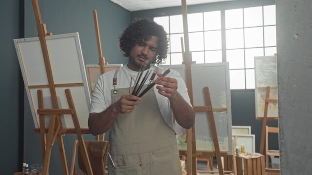 Man painter in studio holds several paintbrushes and inspects a brush tip with bare hand, smiling among easels and canvases; quiet concentration.