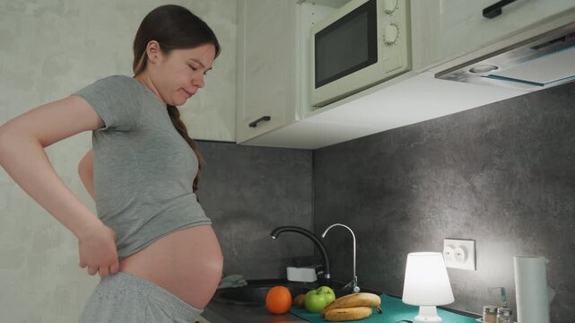 Pregnant caucasian woman cradling belly, adjusting waistband and rubbing lower back, standing by countertop with fruits and lamp, quiet expectant moment, tender connection and mild discomfort