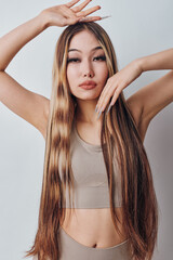 Fototapeta premium Young woman with long straight hair posing with hands near face and head in beige sleeveless top against neutral background for fashion and beauty.