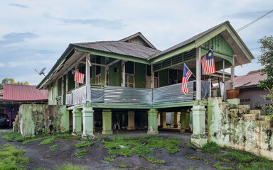 Obraz premium Weathered traditional Malay stilt house with Malaysian flags, Kampong Bharu, Kuala Lumpur, Malaysia. Green wooden structure, corrugated roof, raised on stilts, under a cloudy sky