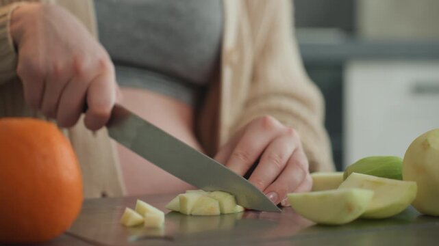 Pregnant woman slicing apple on countertop, quiet morning scene with expectant mother preparing vitaminrich fruit snack, closeup hands chopping apple beside orange, soft natural light, cozy cardigan,