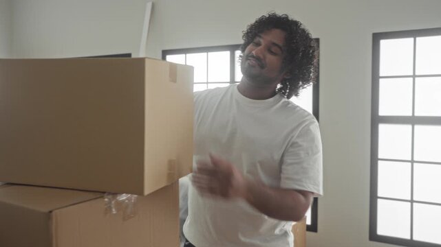Man in white tshirt smiling with arms crossed beside stacked cardboard boxes in a building; pride contentment.