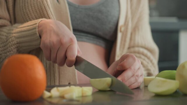 Pregnant woman chopping fruit near belly, playful intimate moment of bonding in warm kitchen, expectant parent in cardigan preparing snack, hands hover over neat slices beside orange, soft light,