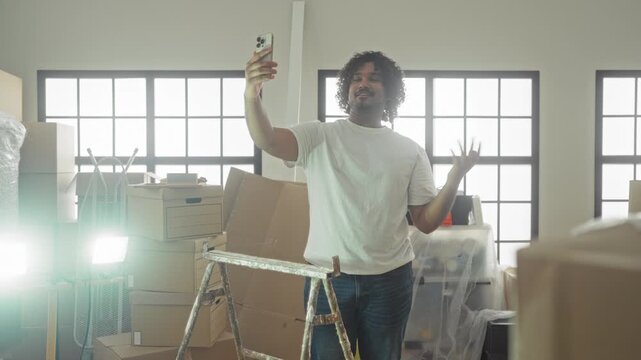 Man holds smartphone for selfie with hand on hip amid packed moving boxes and stepladder in building; new home pride.