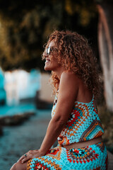 Smiling woman in sunglasses wearing a colorful crochet dress sitting relaxed outdoors at sunset © simona