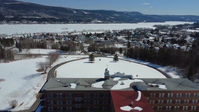 Wide drone shot showcasing the roof and architecture of a former convent in Quebec. This religious heritage site overlooks the village of Lac-au-Saumon, Canada, under a pristine blanket of snow.