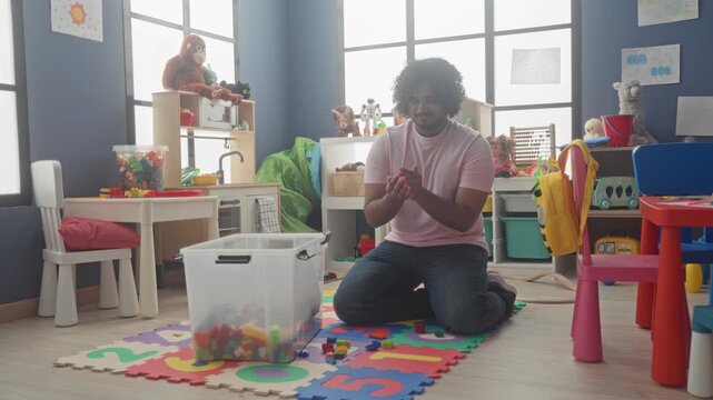 Man kneels sorting colorful toy blocks with hands on a foam play mat in a kindergarten building; playful care.